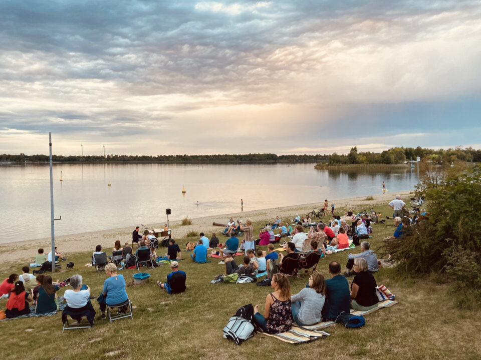Leipziger Tastentage 2025 Mehrere Menschen lauschen auf Decken oder Klappstühlen am Kulkwitzer See dem Konzert von Mr. Pianoman