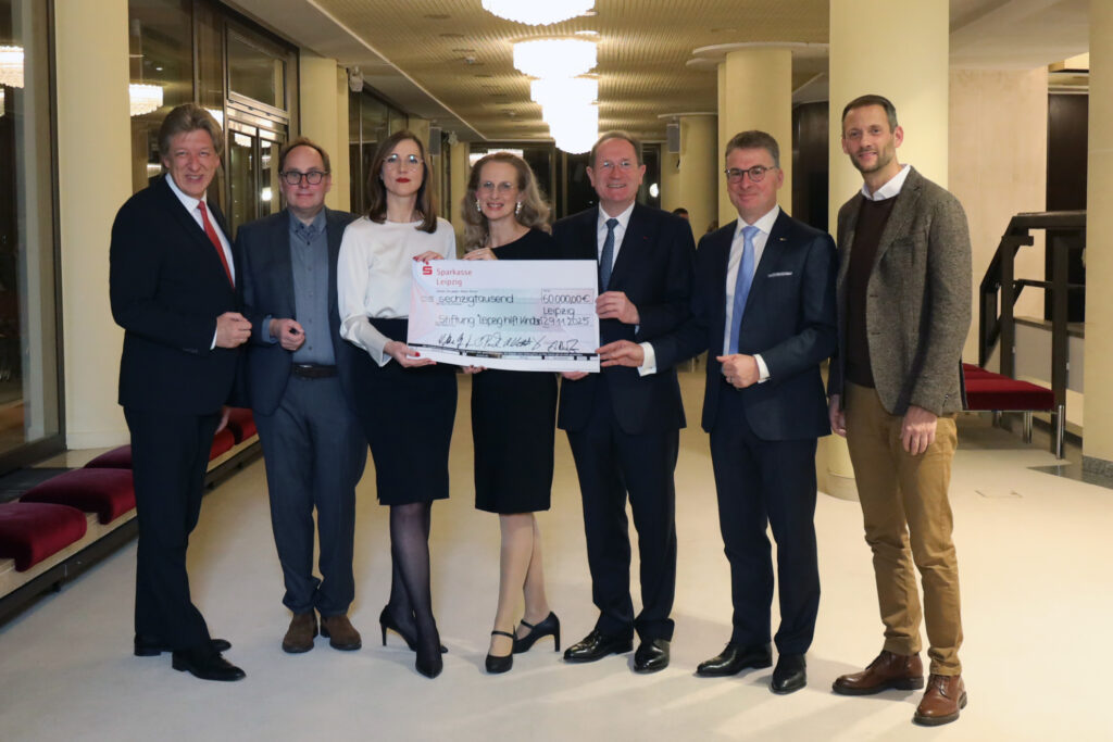 Fünf Männer und zwei Frauen in formeller Kleidung stehen in einem eleganten Flur nebeneinander und blicken in die Kamera. Die zwei Frauen und zwei Männer halten einen symbolischen Spendenscheck in die Kamera.